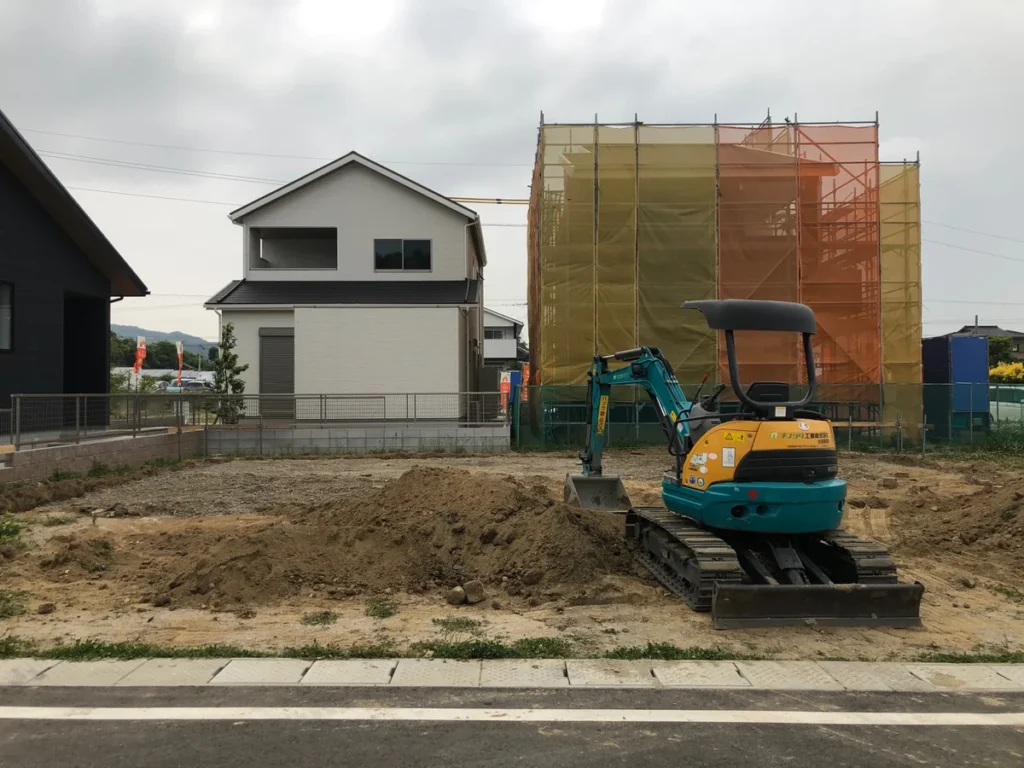 道路側から見た掘削中の敷地と、手前に停まる小型バックホー