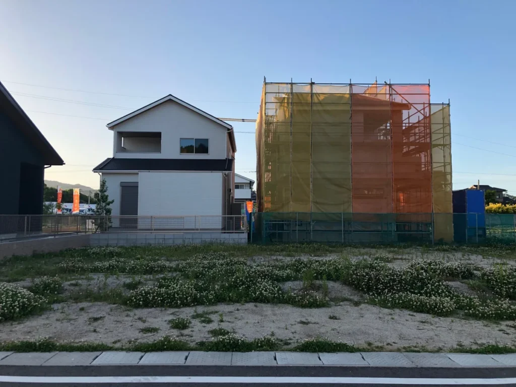 道路側から見た更地の敷地と、左の白い住宅、右の養生ネットで囲まれた建物