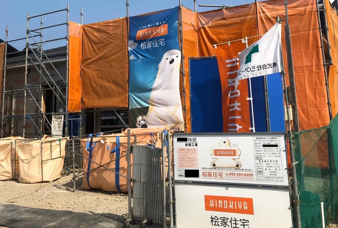 足場とオレンジ色の養生シートで囲われた新築住宅の工事現場外観