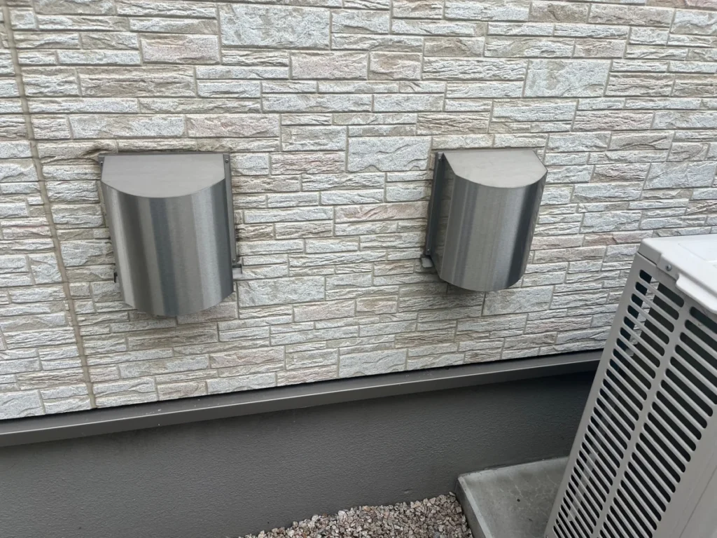 住宅外壁に並ぶ2つの金属製換気フードと室外機の一部