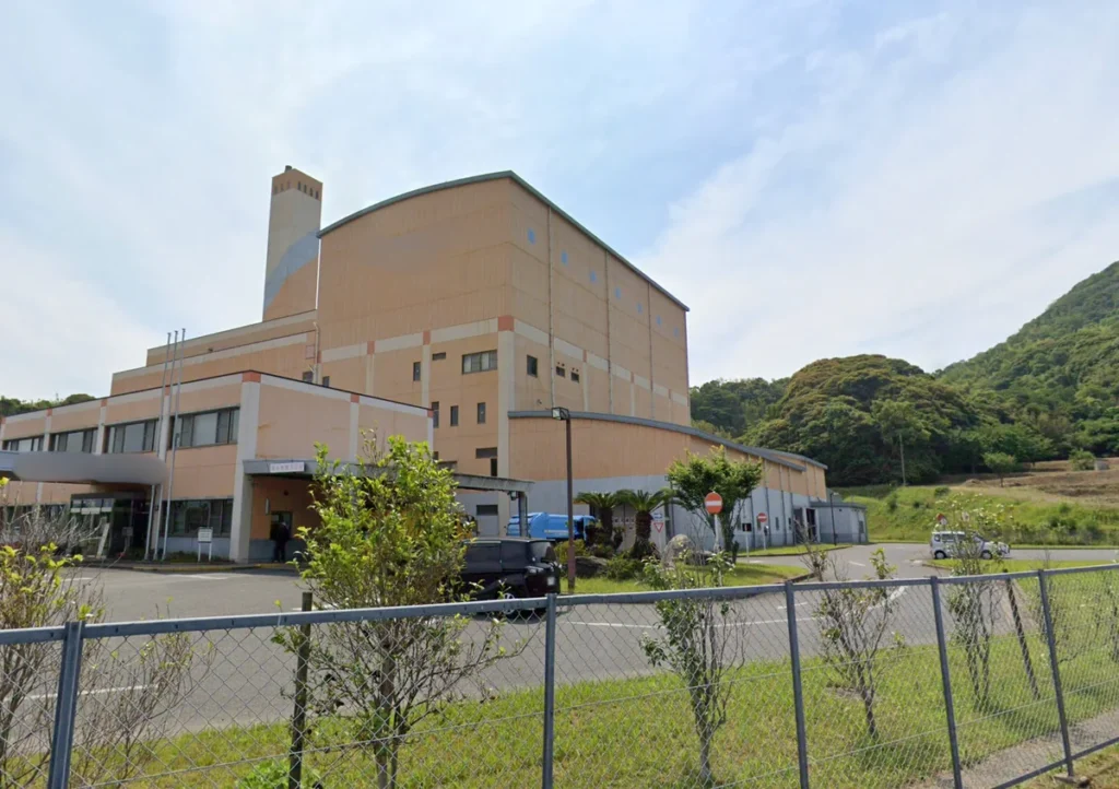 山林を背景に建つクリーンセンターの外観。手前には駐車場とフェンスが見える。