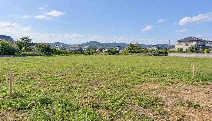 草と土のある平坦な住宅用地に無地の測量杭が立ち、遠くに住宅街と山並みが見える。