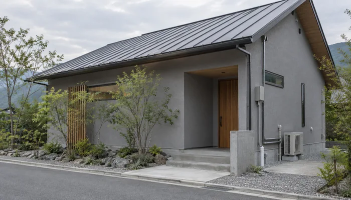 灰色の外壁と金属屋根の戸建てに、雨どいと玄関脇の屋外設備が見える