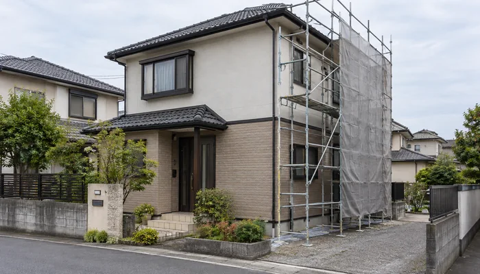 住宅街の戸建ての外壁に足場が組まれ、周囲の住宅と曇り空が見える